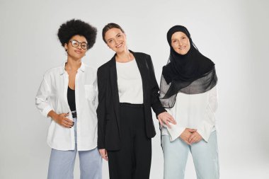 Gri üzerinde yalnız poz verirken kameraya bakan çok ırklı kadınlar gülümsüyor.