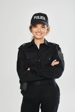 Üniformalı neşeli kadın polis kollarını kavuşturup gri renkte poz veriyor.