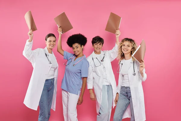 stock image positive interracial doctors holding paper folders isolated on pink, breast cancer awareness