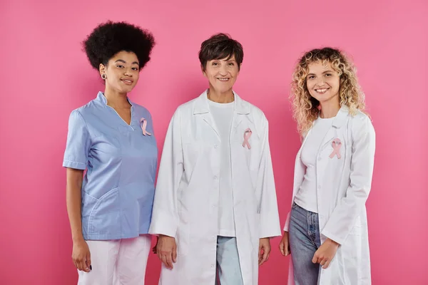 ピンクの背景に立っている乳がんのリボンを持つ前向きな人種間の医師 — ストック写真