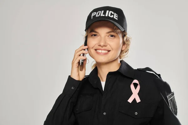 stock image joyful policewoman with pink ribbon of breast cancer talking on smartphone isolated on grey