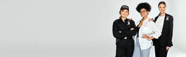 Радостные межрасовые женщины с розовыми лентами рака груди обнимаются изолированно на сером, баннер