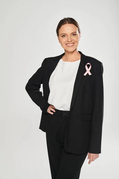 Stock image smiling businesswoman with pink ribbon on jacket isolated on grey, breast cancer awareness