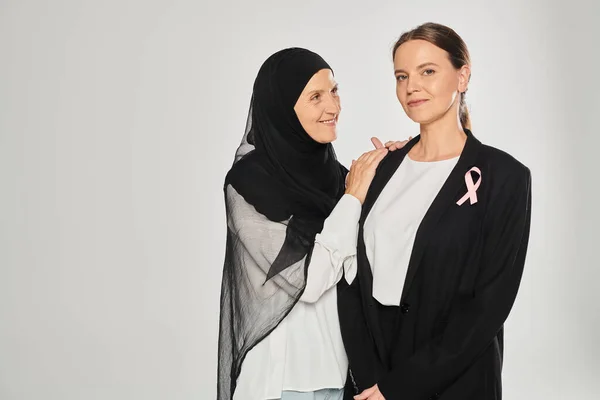 stock image smiling woman in hijab hugging businesswoman with pink ribbon isolated on grey, breast cancer