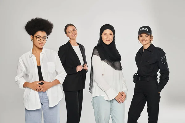 group of multiethnic women looking at camera and smiling while posing isolated on grey