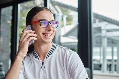 Gözlüklü neşeli genç adam cam arka planda telefonla konuşuyor, iş birliği yapıyor.