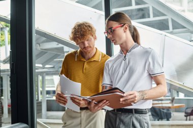 two male colleagues concentrated at working papers near window on backdrop, coworking concept clipart