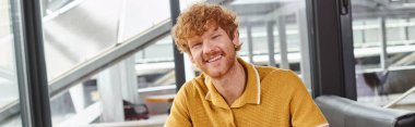 happy red haired man smiling and looking at camera with window backdrop, coworking concept, banner clipart
