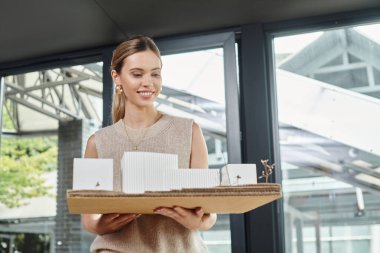 Akıllı pastel giyimli neşeli bir çalışan yapı ve tasarım bürosunun ölçekli modeline bakıyor.