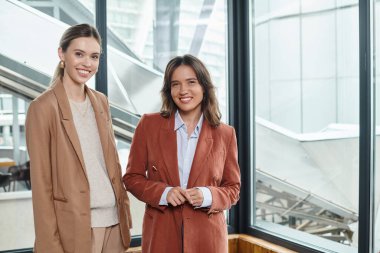 two female colleagues in stylish formal attire posing on window background, coworking concept