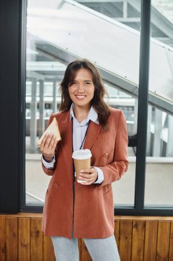 cheerful brunette employee smiling at camera and enjoying her lunch break, coworking concept clipart