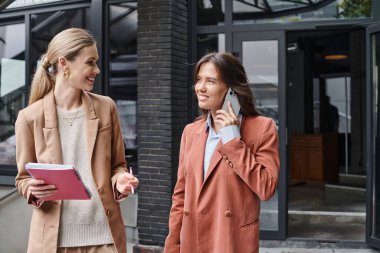 Genç bayan iş arkadaşları ellerinde kağıtlarla dışarıda duruyorlar ve telefonla konuşuyorlar, iş birliği yapıyorlar.