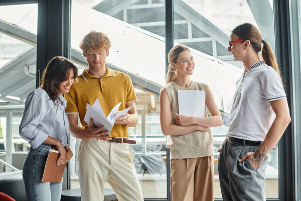 young coworkers in business casual outfits with work papers with glass on backdrop, coworking
