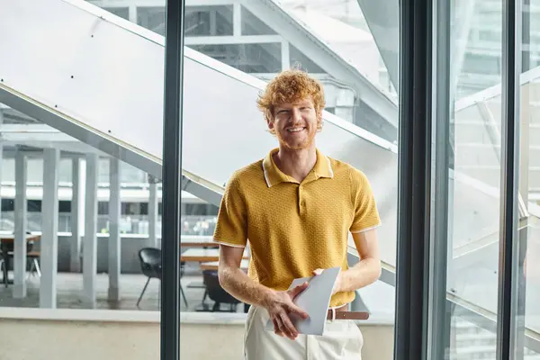 stock image young red haired man in smart casual attire with documents smiling looking at camera, coworking