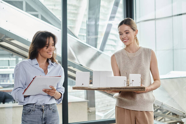 young coworkers examining scale model and making notes with window on backdrop, design bureau