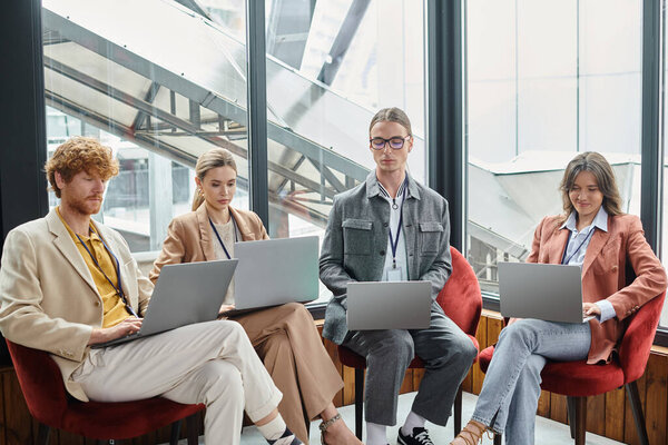 four concentrated team members working on their laptops with window on backdrop, coworking concept