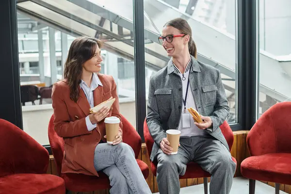 stock image two coworkers in smart casual attire sitting in chairs chatting and eating, coworking concept