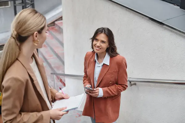 stock image two colleagues standing on stairs looking at each other holding phone and documents, coworking