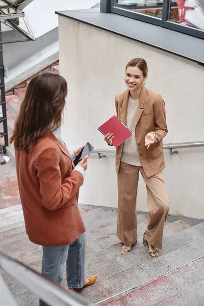 stock image two young colleagues standing on stairs discussing work with phone and papers in hands, coworking