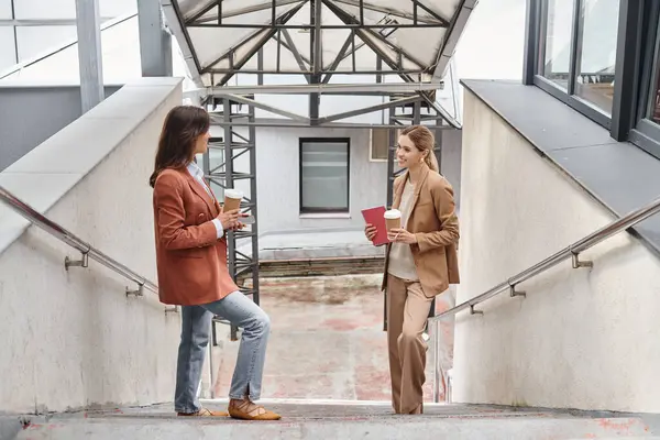 Stock image two colleagues in business casual attire standing on stairs smiling at each other, coworking concept