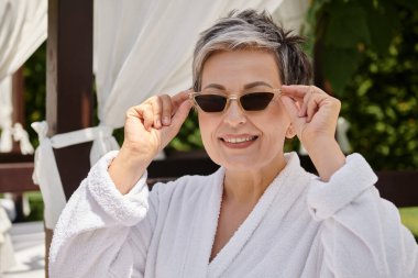 happy middle aged woman wearing sunglasses and looking at camera during wellness retreat on vacation clipart