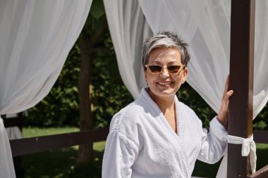 Güneş gözlüklü neşeli olgun kadın özel köşkünde dinleniyor, lüks bir tatil köyünde huzur içinde dinleniyor.