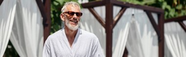 Şık güneş gözlüklü neşeli orta yaşlı adam lüks bir tatil köyünde güneşleniyor.