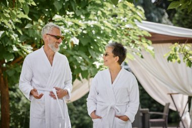 Orta yaşlı bir adam güneş gözlüğü ve bornozlu mutlu eşiyle sohbet ediyor, yaz bahçesi, sağlık inzivası.