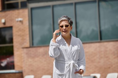 Beyaz cüppeli, güneş gözlüklü orta yaşlı bir kadın lüks bir tatil köyünde havuz başında akıllı telefondan konuşuyor.