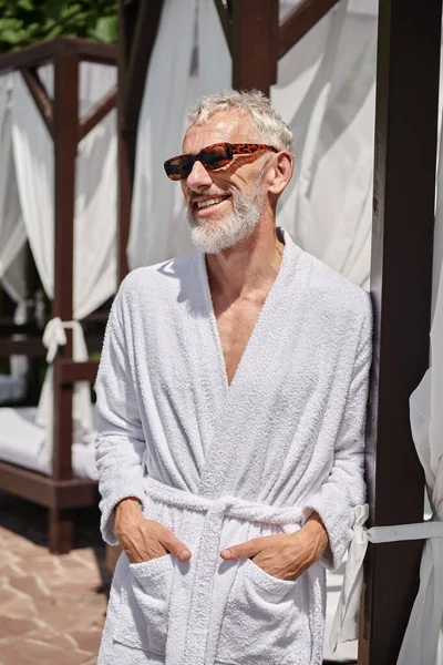 cheerful mature man in sunglasses and robe posing with hands in pockets near private pavilion