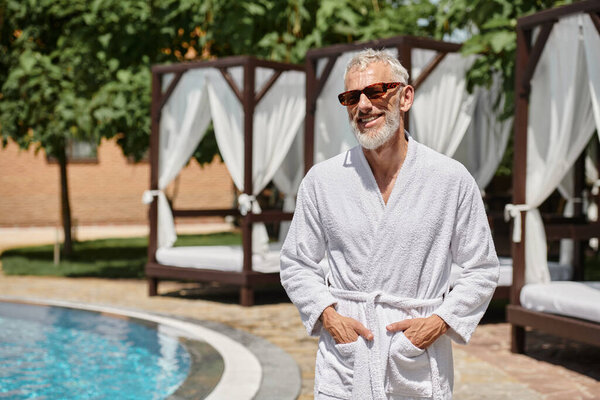 cheerful mature man in sunglasses and robe posing with hands in pockets near poolside at resort