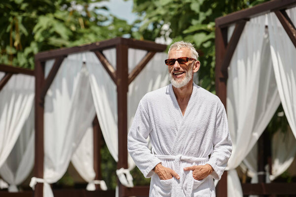 cheerful middle aged man in sunglasses and robe posing with hands in pockets in luxury resort