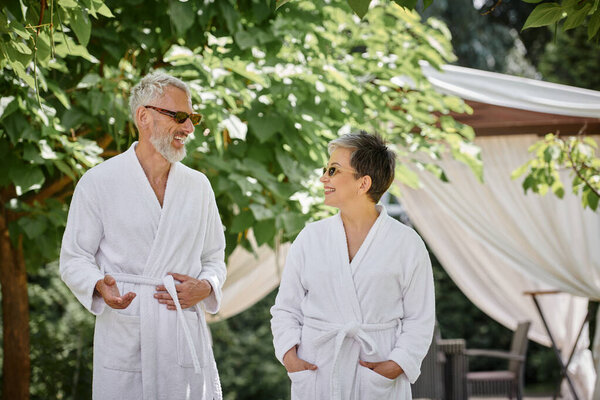 middle aged man chatting with happy wife in sunglasses and robe, summer garden, wellness retreat
