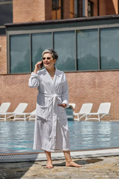 happy middle aged woman in robe and sunglasses talking on smartphone at poolside in luxury resort