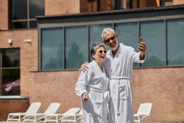 Lüks bir tatil köyünde, kaftanlı mutlu orta yaşlı çift akıllı telefondan selfie çekiyor.