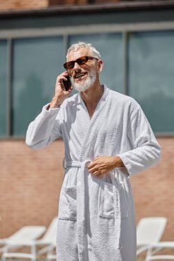 Mutlu ve dövmeli orta yaşlı adam güneş gözlüklü ve bornozlu tatil köyünde akıllı telefonla konuşuyor, geri çekiliyor.