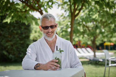 wellness retreat concept, happy middle aged man in sunglasses and robe enjoying cocktail on vacation clipart