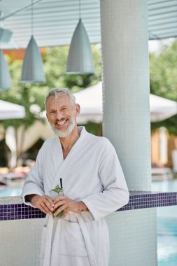 Beyaz cüppeli orta yaşlı adam gülümsüyor ve havuz kenarında mojito kokteyli tutuyor.