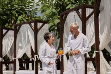 happy mature couple in white robes holding cocktails and talking near pavilion, wellness retreat clipart