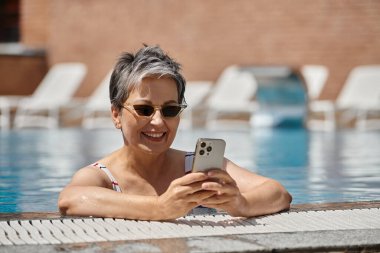 Yüzme havuzunda akıllı telefon kullanan, güneş gözlüklü neşeli orta yaşlı bir kadın.
