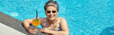happy mature woman in sunglasses swimming in pool with blue water and holding cocktail, banner clipart