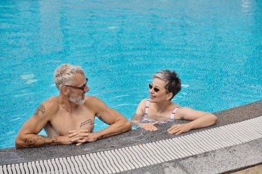 Güneş gözlüklü orta yaşlı çift tatil sırasında havuz içinde sohbet ediyorlar.