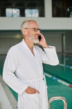 Beyaz cüppeli ve gözlüklü neşeli olgun bir adam kaplıca merkezinde akıllı telefondan konuşuyor, geri çekiliyor.