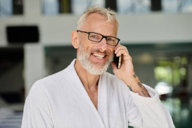 positive mature man in white robe and glasses talking on smartphone inside of indoor spa center clipart