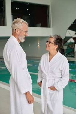 joyful middle aged couple in glasses and white robes standing near indoor pool in spa center clipart