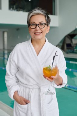 Spa ve geri çekilme konsepti, gözlüklü ve bornozlu mutlu olgun kadın kapalı havuz kenarında kokteyl tutuyor.