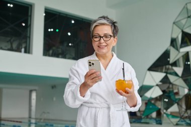 Sağlık ve spa konsepti, elinde kokteyl tutan ve akıllı telefon kullanan mutlu, olgun bir kadın.