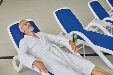 Sağlık konsepti, gözlüklü ve cüppeli olgun bir adam spa merkezindeki kokteylin yanında dinleniyor.