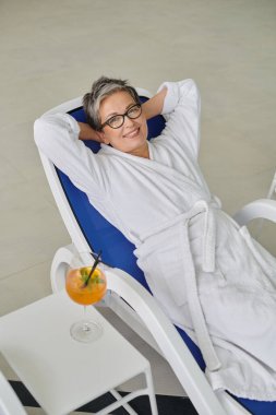 Geri çekilme konsepti, beyaz cüppeli mutlu olgun kadın spa merkezindeki kokteylin yanında dinleniyor.