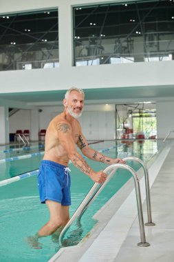 Dövmeli ve sakallı mutlu olgun bir adam bilardo merdiveninin yanındaki mavi suda duruyor.